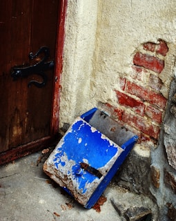 A weathered blue metal mailbox lies on a concrete floor against a wall. The wall has peeling, exposed red bricks and beige plaster. A wooden door with an ornate black hinge is partially visible. Leaves are scattered on the ground around the mailbox.