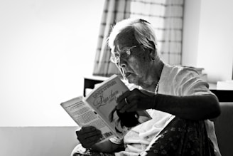 An elderly person with glasses is sitting in a cozy room, deeply engrossed in reading a book. The setting suggests a serene and quiet environment, highlighted by a softly lit window with curtains in the background.