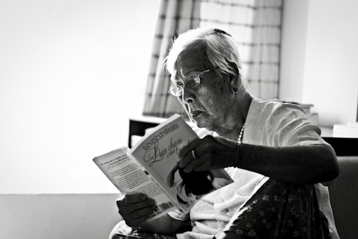 An elderly person with glasses is sitting in a cozy room, deeply engrossed in reading a book. The setting suggests a serene and quiet environment, highlighted by a softly lit window with curtains in the background.