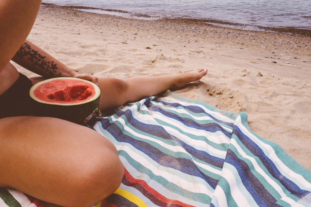 uma pessoa sentada em uma toalha na praia com uma melancia
