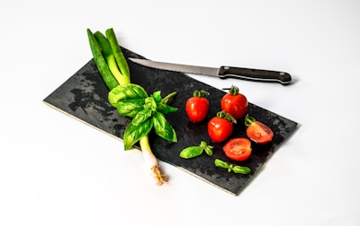 A black cutting board is placed on a white surface, featuring several whole and sliced red tomatoes along with a few fresh green onions and basil leaves. A black-handled kitchen knife is resting on the board beside the vegetables.