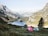 Two colorful tents are set up on a grassy hillside overlooking a serene mountain lake surrounded by tall, rocky mountains. The landscape is lush and green, with patches of trees and vegetation. The sky is clear, enhancing the peacefulness of the scene.