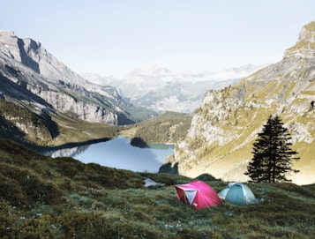 Two colorful tents are set up on a grassy hillside overlooking a serene mountain lake surrounded by tall, rocky mountains. The landscape is lush and green, with patches of trees and vegetation. The sky is clear, enhancing the peacefulness of the scene.