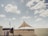 A large triangular shade structure is set in an open, sandy area under a partly cloudy sky. The structure is supported by three poles, with the central pole being the tallest. Surrounding the structure is a wooden fence and some sparse vegetation. There are various objects like a trash can and a small building in the background, creating a semi-deserted feel.