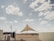 A large triangular shade structure is set in an open, sandy area under a partly cloudy sky. The structure is supported by three poles, with the central pole being the tallest. Surrounding the structure is a wooden fence and some sparse vegetation. There are various objects like a trash can and a small building in the background, creating a semi-deserted feel.