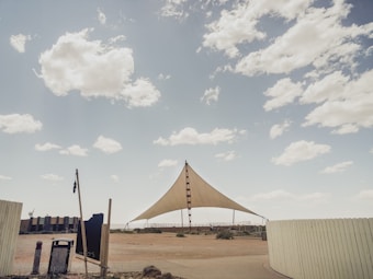 A large triangular shade structure is set in an open, sandy area under a partly cloudy sky. The structure is supported by three poles, with the central pole being the tallest. Surrounding the structure is a wooden fence and some sparse vegetation. There are various objects like a trash can and a small building in the background, creating a semi-deserted feel.