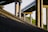 Concrete bridge pillars support an overpass structure, casting shadows against a sloped embankment made of rocks. The play of light and shadow creates geometric patterns, and greenery is visible in the background.