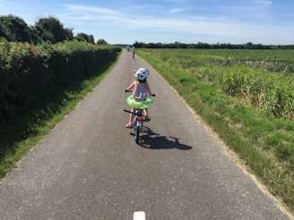 A child wearing a helmet rides a bicycle on a paved path surrounded by lush greenery and open fields. The sky is clear and blue, indicating a sunny day, and the child has a swim ring around their waist. Another cyclist can be seen further down the path.