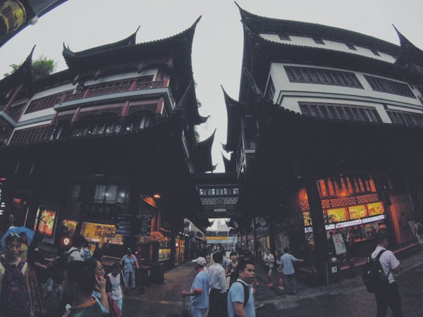 An architectural scene featuring traditional Asian-style buildings with ornate, curved roofs. The structures are tall, with multiple stories and detailed woodwork. The street below is bustling with people, some engaged in conversation, and there are signs and bright lights from commercial establishments.