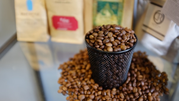 A black metal cup is overflowing with coffee beans, which are also scattered around the base. Behind the cup, several bags of coffee beans are displayed, each with a distinctive label.