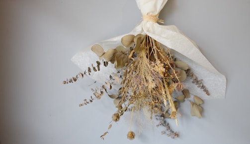 A bouquet of dried flowers wrapped in white paper, featuring various types of dry foliage and plants arranged in an artistic manner. The bouquet is hanging against a plain, light gray wall.
