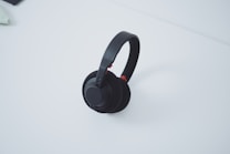 A pair of black over-ear headphones resting on a white surface. The headphones have cushiony ear pads and a sleek headband with a minimalist design.