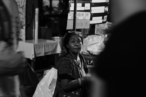 A middle-aged woman is sitting near a food stall, surrounded by signs and packages. The scene is captured in black and white, highlighting the focus on the woman who appears thoughtful or pensive as she gazes into the distance.