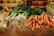 A farmer's market stall displaying a variety of fresh vegetables, including leeks with long roots and a bunch of bright orange carrots. The produce is arranged neatly on a burlap-covered table, surrounded by wooden crates. The background includes more vegetables and small chalkboard signs.