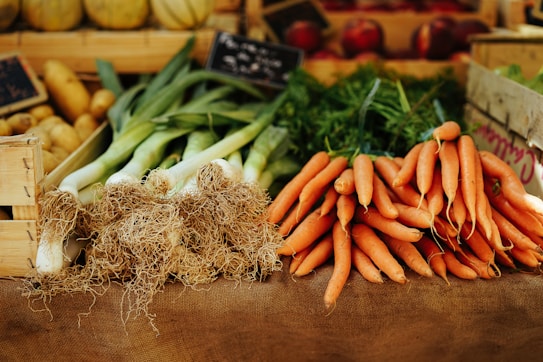 A farmer's market stall displaying a variety of fresh vegetables, including leeks with long roots and a bunch of bright orange carrots. The produce is arranged neatly on a burlap-covered table, surrounded by wooden crates. The background includes more vegetables and small chalkboard signs.