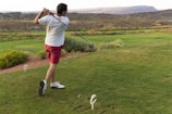 A man swings a golf club on a grassy course, wearing a white shirt, red shorts, and white shoes. The landscape features rolling hills with patches of shrubs and bushes. In the distance, there are power lines and a mountainous backdrop under an overcast sky.