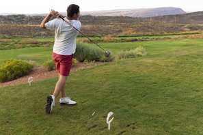 A man swings a golf club on a grassy course, wearing a white shirt, red shorts, and white shoes. The landscape features rolling hills with patches of shrubs and bushes. In the distance, there are power lines and a mountainous backdrop under an overcast sky.
