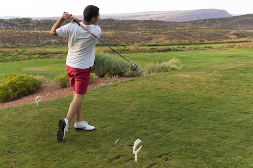 A man swings a golf club on a grassy course, wearing a white shirt, red shorts, and white shoes. The landscape features rolling hills with patches of shrubs and bushes. In the distance, there are power lines and a mountainous backdrop under an overcast sky.
