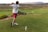 A man swings a golf club on a grassy course, wearing a white shirt, red shorts, and white shoes. The landscape features rolling hills with patches of shrubs and bushes. In the distance, there are power lines and a mountainous backdrop under an overcast sky.