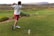 A man swings a golf club on a grassy course, wearing a white shirt, red shorts, and white shoes. The landscape features rolling hills with patches of shrubs and bushes. In the distance, there are power lines and a mountainous backdrop under an overcast sky.