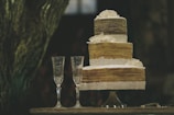 A rustic, three-tiered cake adorned with lace and white flowers stands on a table. Two crystal champagne flutes accompany the cake, and a silver cake server lies nearby.