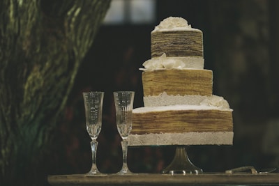 A rustic, three-tiered cake adorned with lace and white flowers stands on a table. Two crystal champagne flutes accompany the cake, and a silver cake server lies nearby.