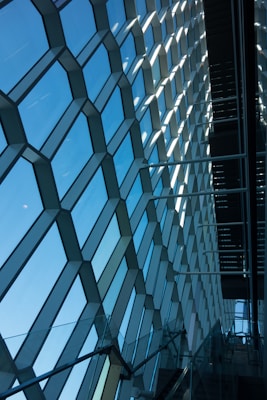 A geometric pattern of large hexagonal glass panels creating a wall, allowing natural light to flood the interior. The panes are framed in dark metal, and a staircase with a glass railing is visible in the lower part, adding depth and perspective to the scene. The design reflects modern architectural aesthetics and connects the interior with the sky outside.