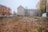 An urban scene featuring an empty lot with sparse vegetation and dry leaves covering the ground. Surrounding the lot are mid-sized residential buildings with a few trees in autumn foliage. The sky appears overcast, creating a subdued atmosphere.