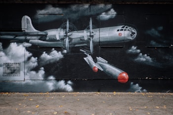 A street mural depicting a World War II-era bomber plane with multiple propellers and two bombs dropping. The artwork is painted in mostly monochromatic tones with contrasting red details on the airplane and bombs. The background includes stylized clouds, adding a dramatic effect to the composition.