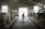 A barn interior featuring several cows tethered in stalls, with farmhands attending to them. The barn has concrete walls, a high ceiling, and a large opening at one end allowing natural light to enter. A farmer holding equipment walks along the center aisle lined with straw.