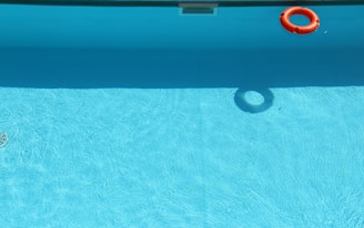 A swimming pool is filled with clear blue water, with a bright orange lifebuoy floating near the edge. The bottom of the pool is visible, and the water is shimmering with sunlight reflections.