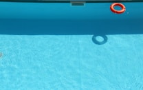 A swimming pool is filled with clear blue water, with a bright orange lifebuoy floating near the edge. The bottom of the pool is visible, and the water is shimmering with sunlight reflections.