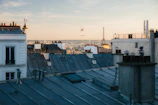 A view over Parisian rooftops with the Eiffel Tower visible in the distance. The scene captures classic chimney stacks and TV antennas under a soft, warm sky during what appears to be late afternoon or early evening.