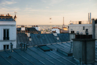 A view over Parisian rooftops with the Eiffel Tower visible in the distance. The scene captures classic chimney stacks and TV antennas under a soft, warm sky during what appears to be late afternoon or early evening.
