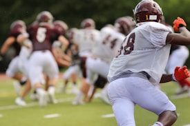 A group of football players wearing maroon and white uniforms are on a field. One player in the foreground, wearing a white jersey and red gloves, is in focus, while more players are visible in the background running during a game or practice.