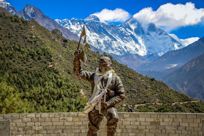A statue of a mountaineer wearing climbing gear stands prominently in the foreground, holding a flag in one hand and a climbing tool in the other. The background features a majestic view of snow-capped mountains and a clear blue sky, with a rugged, green hillside leading up to the peaks.