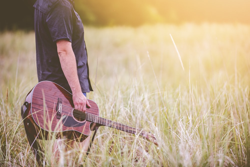 Hombre que lleva la guitarra acústica marrón recortada de pie en el campo de hierba verde