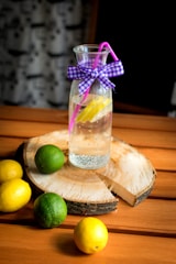 A glass pitcher filled with water and a slice of lemon inside is placed on a wooden slab. The pitcher is adorned with a purple ribbon bow and has a pink straw. Surrounding the wooden slab are whole lemons and limes on a wooden table.