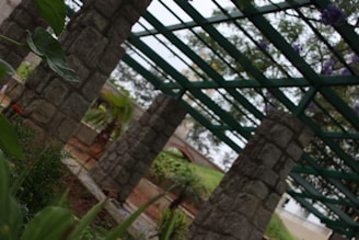 A series of robust stone columns support a green metal framework, resembling a pergola, under a cloudy sky. Lush green foliage and a variety of plants thrive in the foreground and background, adding vibrancy to the structured setting. The scene includes a small curved bridge, partially visible in the background, that suggests a garden or park setting.