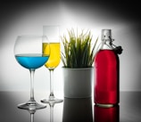 Two glasses, one with blue liquid and the other with yellow liquid, sit to the left of a white potted plant containing green grass-like foliage. To the right, a glass bottle filled with red liquid is placed, all set against a reflective surface and a gradient background.