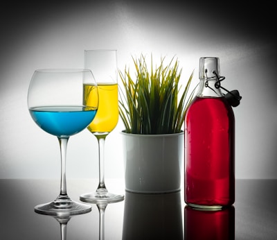 Two glasses, one with blue liquid and the other with yellow liquid, sit to the left of a white potted plant containing green grass-like foliage. To the right, a glass bottle filled with red liquid is placed, all set against a reflective surface and a gradient background.