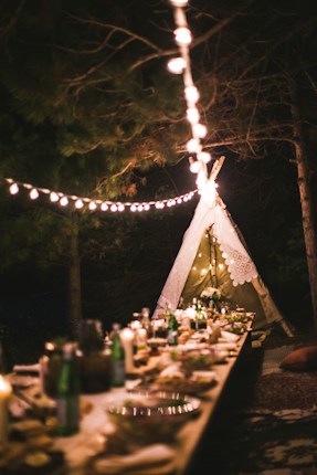 A beautifully set outdoor dinner table is adorned with various dishes, drinks, and decorations. A warm glow from string lights creates a cozy atmosphere, and a decorative teepee tent is lit in the background. The setting is surrounded by trees, adding an enchanting, rustic vibe to the scene.