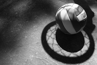A black and white volleyball rests on a textured concrete surface, casting a shadow that forms a circular pattern with distinct outlines. The lighting emphasizes the ball's panels and the surrounding shadow.
