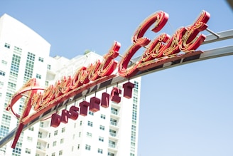 A large, vibrant neon sign reads 'Fremont East District' in cursive style, situated against the backdrop of tall, modern white buildings with abundant windows. The sky appears clear and blue.