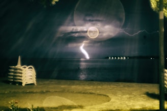 A dark, moody night scene featuring a lightning strike over a body of water. The foreground includes a stack of white plastic chairs. Light reflections and circular lens flares are visible, adding to the mysterious ambiance. Bright spots on the horizon suggest distant lights or buildings.