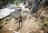 A person herds a group of goats up a dusty slope near a farm. The scene is set in rural surroundings with a building, trees, and various farm equipment in the background. The person is wearing a green hat and is guiding the animals with a stick as they navigate the earthy terrain.