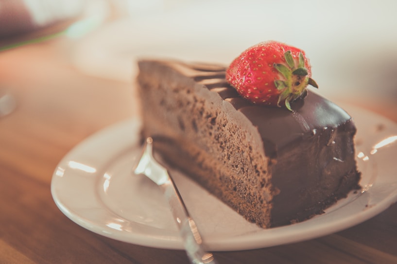 A slice of chocolate cake topped with a fresh strawberry sits on a white plate. The cake has a glossy chocolate glaze, and a small fork is placed beside it. The background is softly blurred, contributing to a warm and inviting atmosphere.