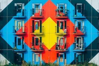 An apartment building facade is adorned with a striking geometric mural composed of bold, vibrant colors such as red, blue, and yellow. Each balcony has black railings, some with potted plants and outdoor furniture, suggesting personal touches by the residents.