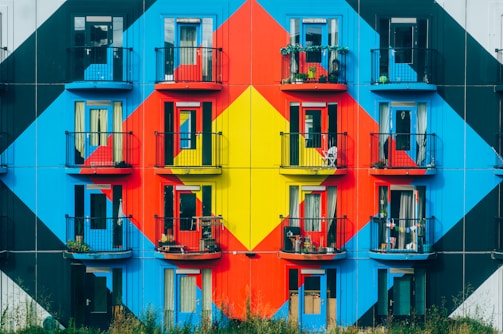 An apartment building facade is adorned with a striking geometric mural composed of bold, vibrant colors such as red, blue, and yellow. Each balcony has black railings, some with potted plants and outdoor furniture, suggesting personal touches by the residents.