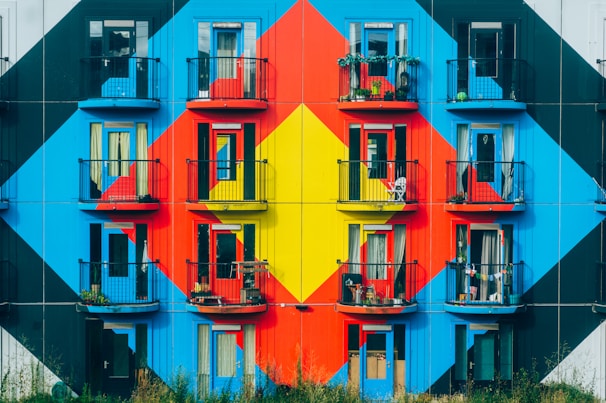 An apartment building facade is adorned with a striking geometric mural composed of bold, vibrant colors such as red, blue, and yellow. Each balcony has black railings, some with potted plants and outdoor furniture, suggesting personal touches by the residents.